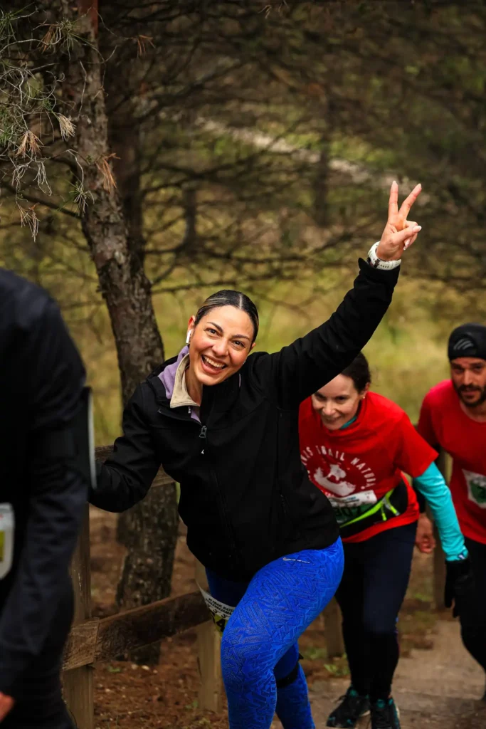 Una mujer con chaqueta negra y mallas azules sonríe y hace el signo de la paz mientras corre cuesta arriba por el Rivas Trail Natura, con otros corredores con camisetas rojas siguiéndola por el bosque tras una ligera lluvia.