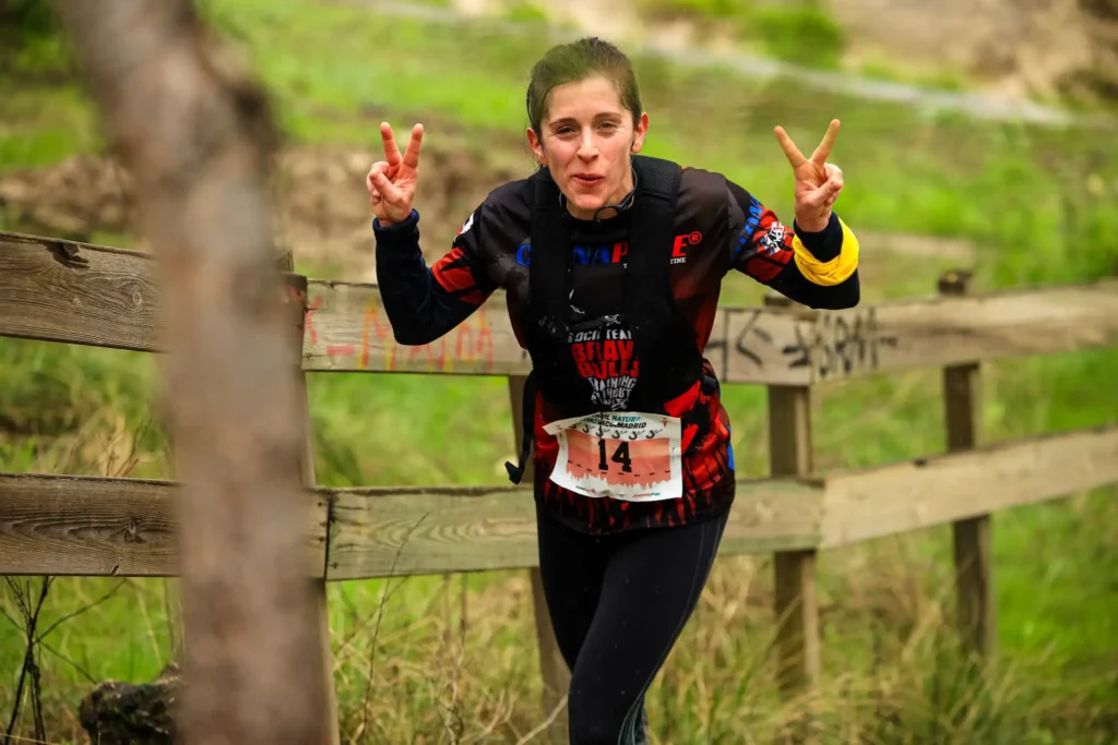 Una mujer corriendo al aire libre por el Rivas Trail Natura, con ropa deportiva y un dorsal con el número 14, levanta ambas manos haciendo signos de paz. En el fondo, una valla de madera y vegetación, captando el espíritu de los corredores.