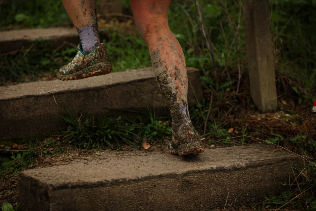 Una persona con las piernas y los zapatos cubiertos de barro sube escalones de hormigón al aire libre en el Rivas Trail Natura, rodeado de hierba y tierra, captando el espíritu de los corredores que desafían los elementos naturales tras una refrescante lluvia.