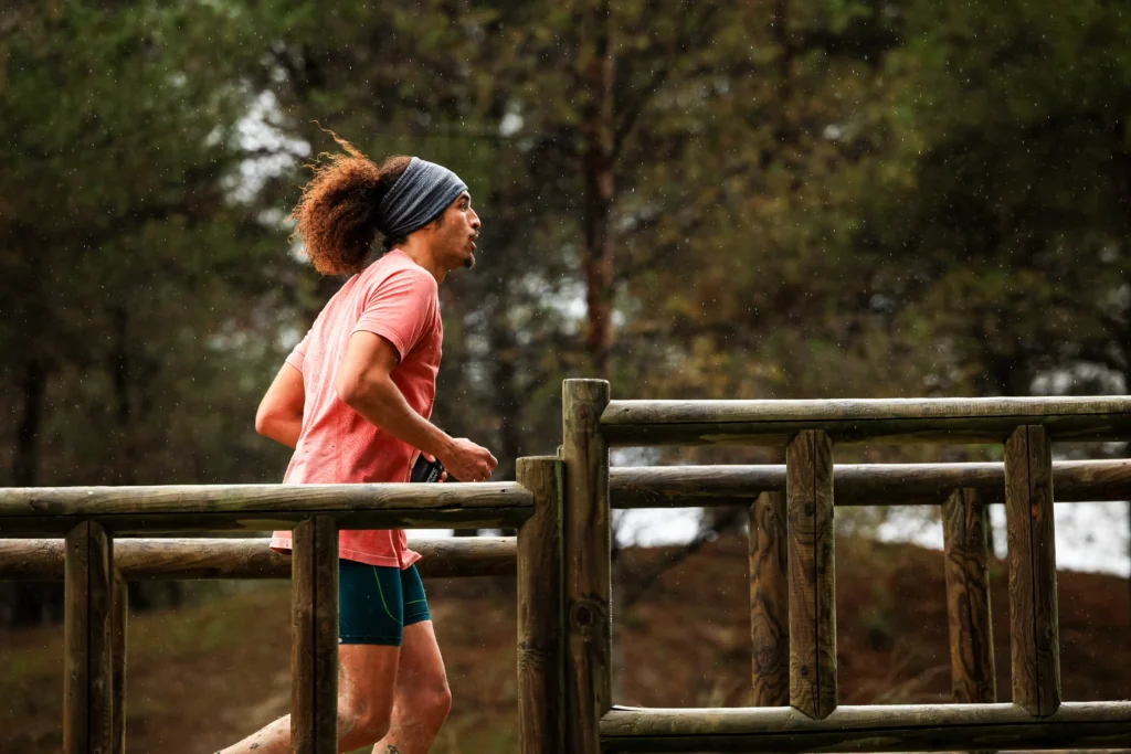 Una persona con camiseta rosa, pantalón corto azul y cinta en la cabeza corre al aire libre por el puente de madera Rivas Trail Natura, con árboles al fondo, uniéndose a otros corredores mientras empieza a caer una ligera lluvia.
