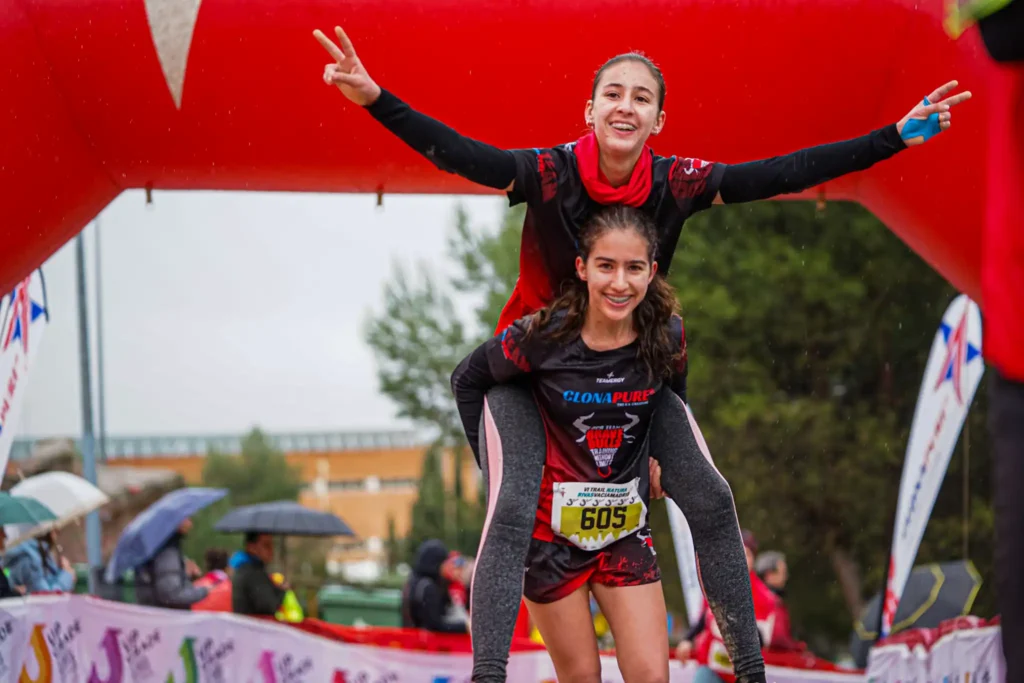 Two young corredores in uniformes de carrera celebran en la meta; uno le da un paseo a caballito al otro, que sonríe y hace signos de paz, bajo el arco hinchable rojo con espectadores y logos de Rivas Trail Natura al fondo.