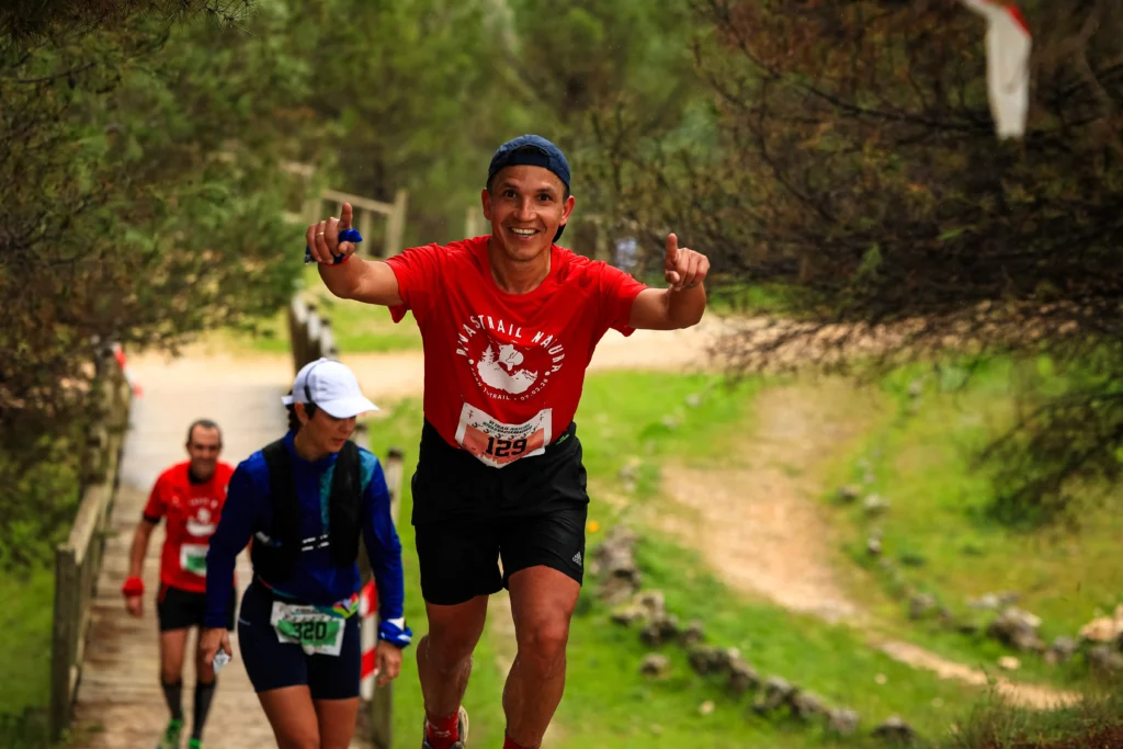 Un corredor con camiseta roja y gorra azul sonríe y gesticula con ambas manos mientras corre por el Rivas Trail Natura, con dos compañeros corredores detrás. La escena al aire libre muestra una exuberante vegetación, un camino de tierra y toques de lluvia reciente.