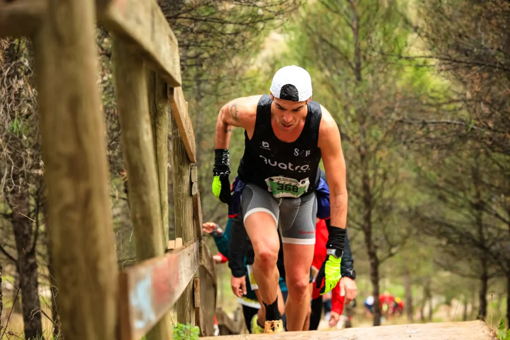 Un corredor con gorra blanca, camiseta de tirantes negra, guantes y dorsal 368 sube bajo la lluvia por un empinado sendero arbolado de Rivas Trail Natura, agarrado a una barandilla de madera mientras otros corredores le siguen entre los árboles.
