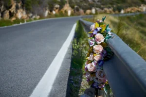 Un ramo de flores artificiales está sujeto a una barandilla metálica junto a una carretera desierta y sinuosa -quizá un monumento silencioso a las tumbas accidentadas- con árboles y terreno rocoso de fondo bajo un cielo despejado.