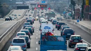Varios carriles de coches y camiones soportan un intenso tráfico durante la operación salida en una autopista. Señales electrónicas en lo alto muestran mensajes, mientras que barreras de hormigón separan los sentidos de circulación. Edificios y árboles se alinean a los lados de la carretera.
