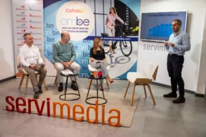 Cuatro personas se encuentran en una moderna sala de conferencias. Tres están sentadas con micrófonos, mientras que una está de pie y presenta un gráfico con los precios medios de las bicicletas. Detrás de ellos se ven logotipos y pancartas promocionales en las paredes.
