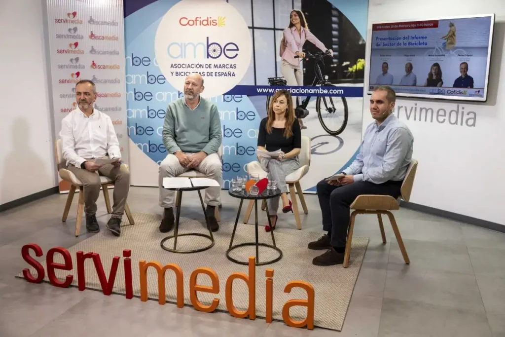Cuatro personas sentadas en sillas en un estudio en el que se ve la marca Cofidis, ambe, Servimedia y Borrador automático. Miran a la cámara, con una pantalla y material promocional sobre bicicletas al fondo.