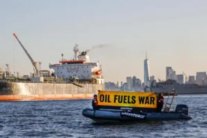 Un pequeño barco de Greenpeace con tres personas sostiene una gran pancarta amarilla en la que se lee OIL FUELS WAR en el agua cerca de una gran nave industrial, destacando cómo los combustibles fósiles impulsan la espiral climática, con el horizonte de una ciudad al fondo.