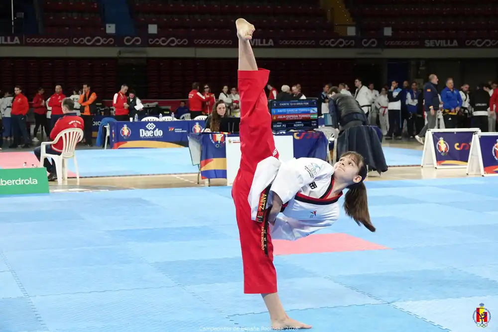 Un artista marcial con uniforme blanco y rojo ejecuta una patada alta sobre una colchoneta en una pista cubierta, con jueces y otros participantes visibles al fondo.