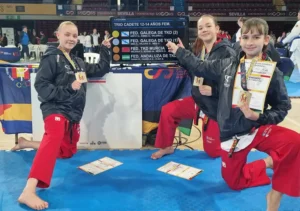 Tres jóvenes artistas marciales vestidas con pantalones rojos y chaquetas negras se arrodillan sobre una colchoneta azul, sosteniendo medallas y certificados, delante de un marcador de competición en el que figuran los equipos y los resultados. Al fondo, otros competidores y jueces.