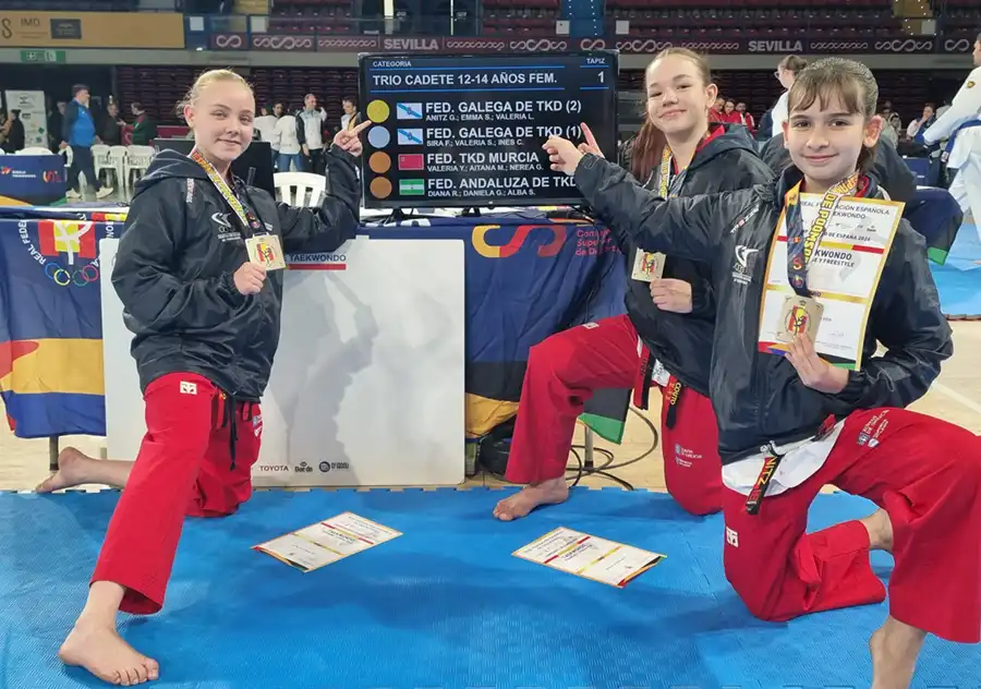 Tres jóvenes artistas marciales vestidas con pantalones rojos y chaquetas negras se arrodillan sobre una colchoneta azul, sosteniendo medallas y certificados, delante de un marcador de competición en el que figuran los equipos y los resultados. Al fondo, otros competidores y jueces.