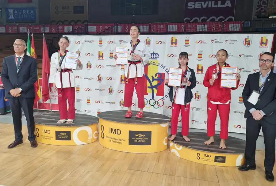 Cuatro jóvenes atletas suben a un podio con diplomas y medallas en un acontecimiento deportivo en pista cubierta, con dos hombres trajeados a cada lado. Detrás de ellos se ve una pancarta con logotipos y texto.