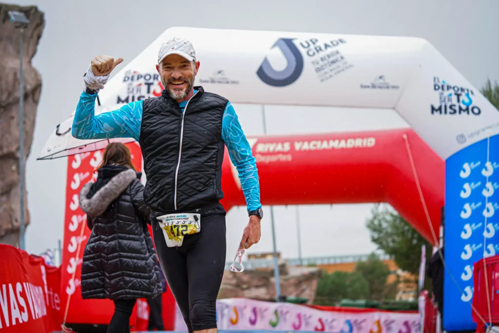 Un corredor vestido con chaleco negro, mangas azules y gorra blanca sonríe y levanta el pulgar mientras cruza la línea de meta de la carrera Rivas Trail Natura con arcos rojos y blancos y pancartas de fondo.