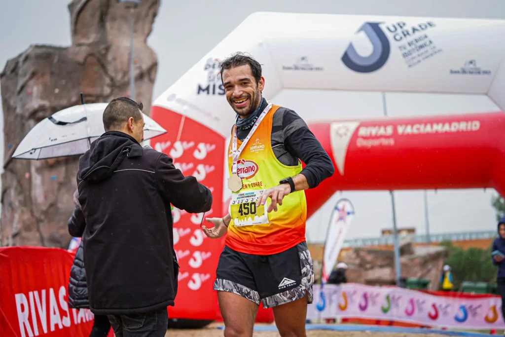 Un corredor con camiseta roja y amarilla y dorsal sonríe y estrecha la mano de otra persona en la lluviosa meta de Rivas Trail Natura, con hinchables, señalización y compañeros corredores al fondo.