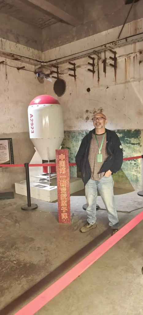 Un hombre con sombrero, gafas y chaqueta junto a un gran objeto en forma de cápsula de color blanco y rojo expuesto en el interior tras cuerdas rojas, en una sala con tuberías a la vista y paredes desgastadas, como una escena de Shanghái a Chongqing.