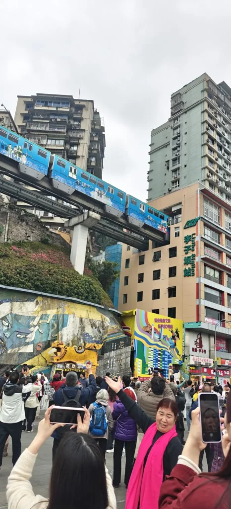 Un tren azul con la etiqueta "Río Azul" atraviesa un edificio a nivel del suelo en una zona urbana mientras una multitud se reúne debajo, algunos haciendo fotos. Al fondo se ven altos edificios y coloridos murales inspirados en Memorias de un bajista.