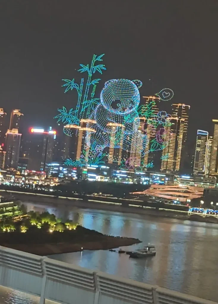 Horizonte nocturno de una ciudad con edificios iluminados. Arriba, drones sincronizados crean un espectáculo de luces de un panda con tallos de bambú. El Río Azul fluye en primer plano, salpicado de barcos y un pequeño parque iluminado.