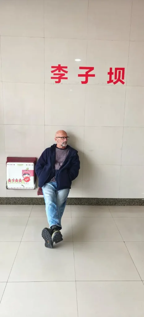 Un hombre calvo, con gafas y barba, quizá un bajista afortunado, se sienta en un banco de pared contra muros de azulejos. Grandes caracteres chinos rojos se elevan sobre él, evocando viajes desde Shanghái a Chongqing. El suelo es de baldosas claras.
