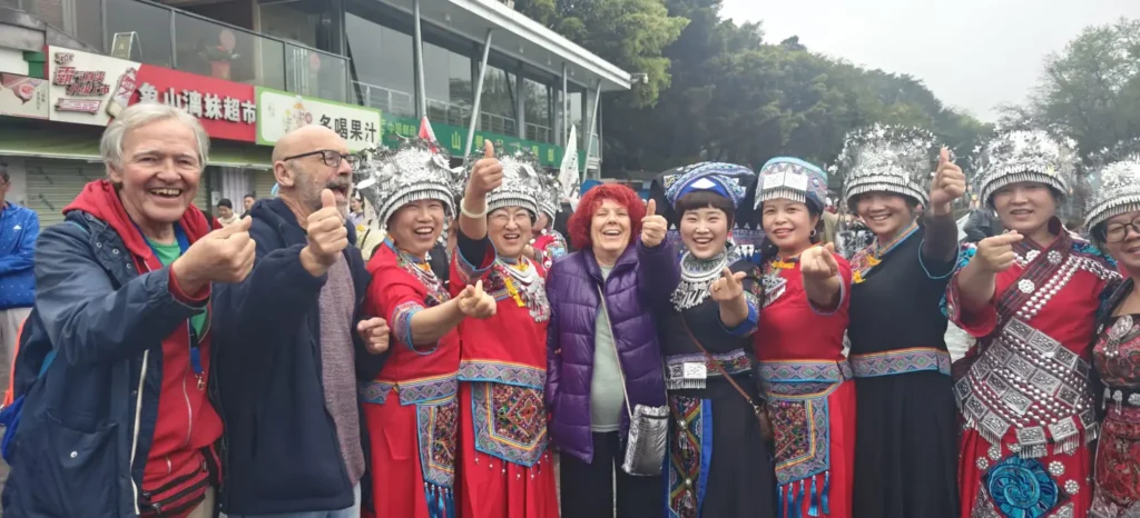 Un grupo de personas, entre ellas turistas y mujeres vestidas con atuendos tradicionales de la etnia china y elaborados tocados plateados, posan juntas al aire libre en Chongqing, sonriendo y haciendo gestos con el pulgar hacia arriba. Al fondo aparecen árboles y un edificio.