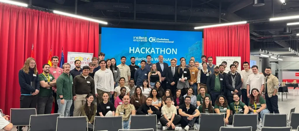 Un gran grupo de personas posan juntas delante de una pantalla que muestra HACKATHON y otros textos en un espacio para eventos de Madrid, con cortinas rojas a ambos lados. La mayoría están de pie mientras que algunos están sentados delante, celebrando las innovaciones de la Smart City.
