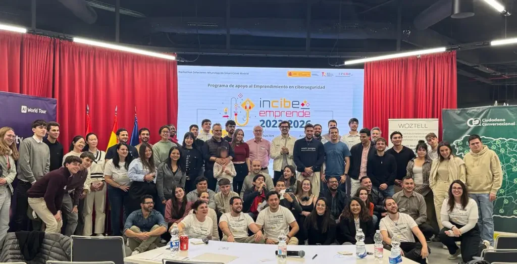 Un nutrido grupo de personas posa para una foto delante de una pantalla que muestra incibe emprende 2022-2023, con banderolas de colores y cortinas rojas de fondo en Madrid. La mayoría sonríe y mira a la cámara en el evento Smart City Conversacional.