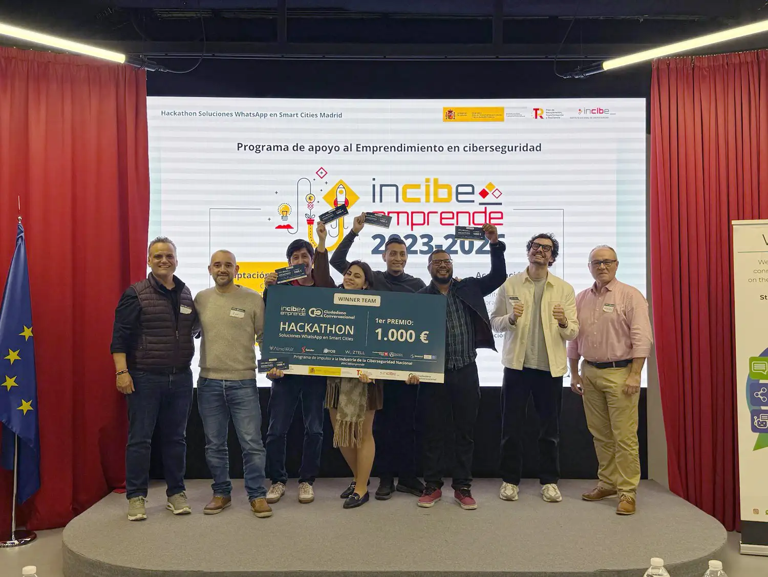 Ocho personas de pie en un escenario de Madrid sostienen un gran cheque de 1.000 euros frente a una pantalla que muestra incibe emprende 2023-2024. Hay banderas, pancartas y un telón rojo en este acto temático sobre la Smart City. Ocho personas de pie en un escenario de Madrid sostienen un gran cheque de 1.000 euros frente a una pantalla que muestra incibe emprende 2023-2024. Hay banderas, pancartas y un telón rojo en este acto temático sobre la Smart City.