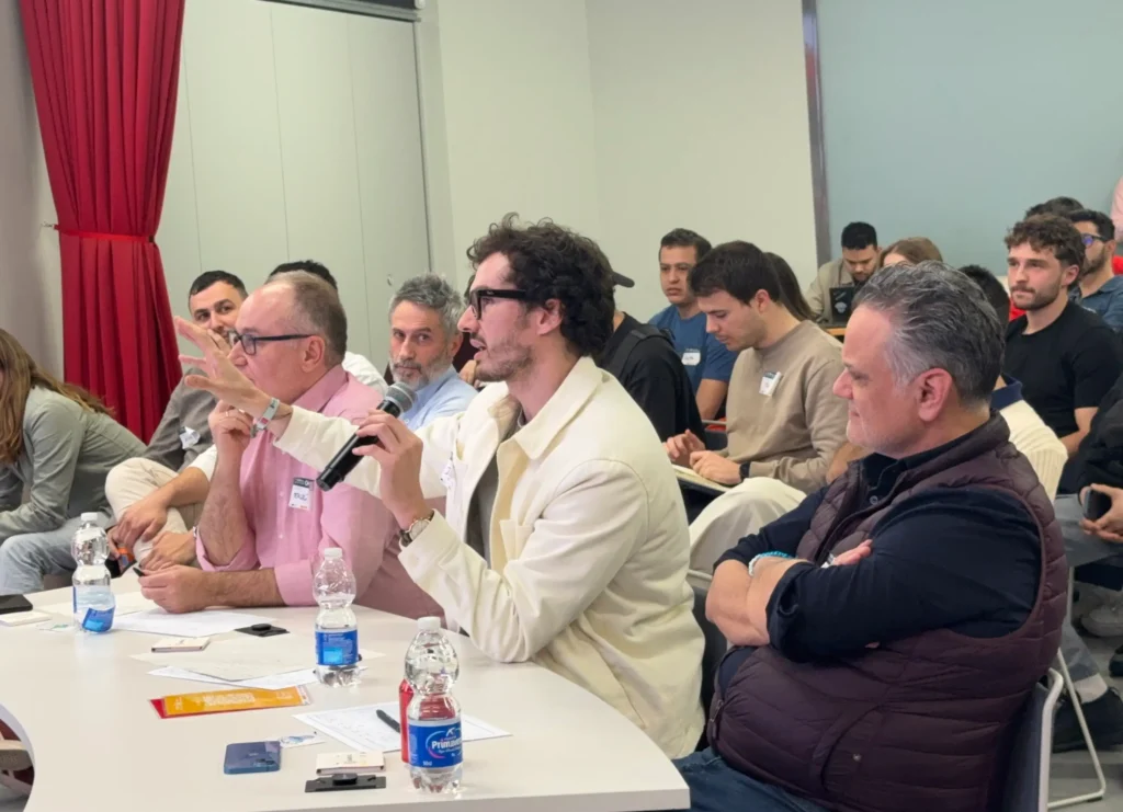 Un grupo de personas sentadas a una mesa en una sala muy iluminada de Madrid, escuchan y toman notas. Una persona en primer plano sostiene un micrófono, mientras otros tienen agua embotellada y papeles, debatiendo sobre innovaciones de Smart City.