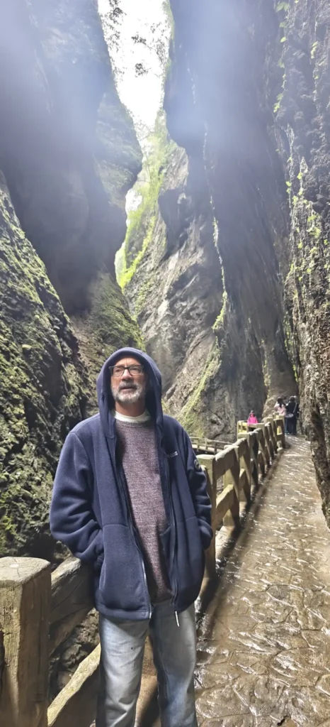 Un hombre vestido con una sudadera azul se encuentra en un estrecho sendero entre altas paredes rocosas cubiertas de musgo en un cañón que recuerda al Río Azul. En el fondo se ve a otras personas a lo largo del sendero, y el cielo es parcialmente visible desde arriba.