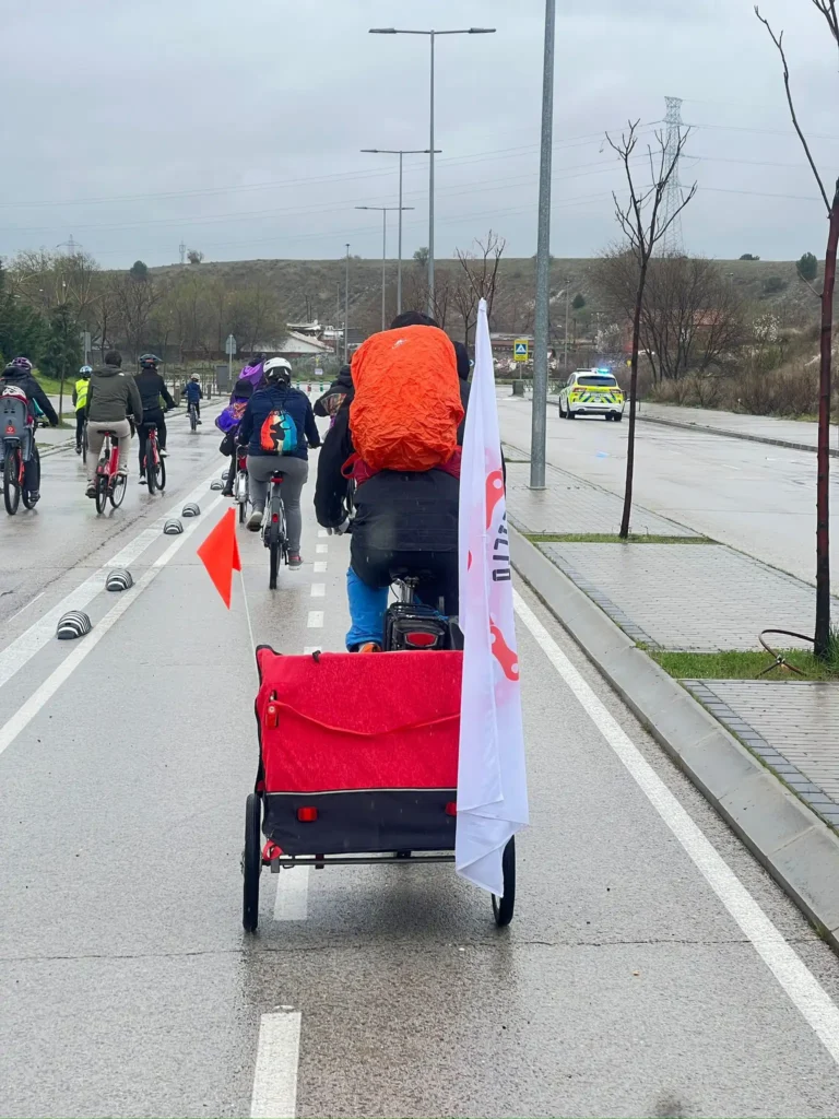 Un grupo de ciclistas circula en bicicleta por una vía urbana mojada de Rivas durante la Bicicletada 7M, con una bicicleta tirando de un remolque rojo para niños que exhibe una bandera blanca. Los ciclistas llevan cascos y mochilas mientras se ve un coche de policía al fondo.