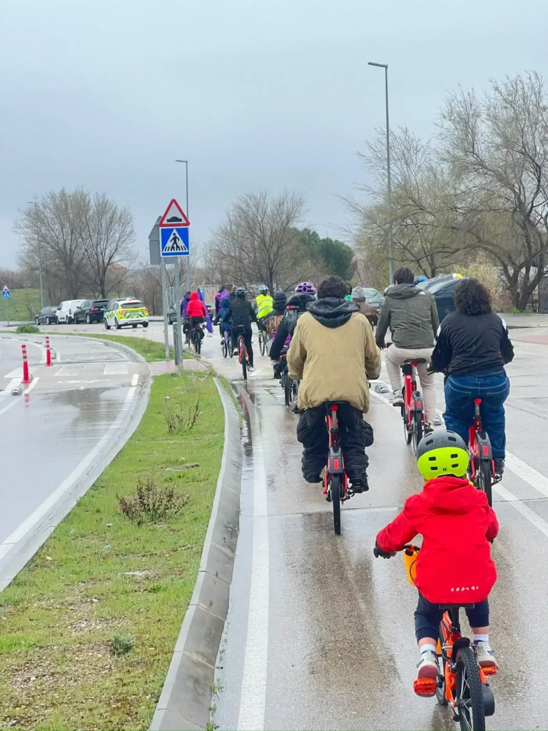 Un grupo de ciclistas, entre ellos niños y adultos, montan en bicicleta por una carretera mojada bajo un cielo nublado durante la Bicicletada 7M en Rivas. Un niño con chaqueta roja y casco verde sigue al grupo entre árboles y señales de tráfico a lo largo de la ruta.