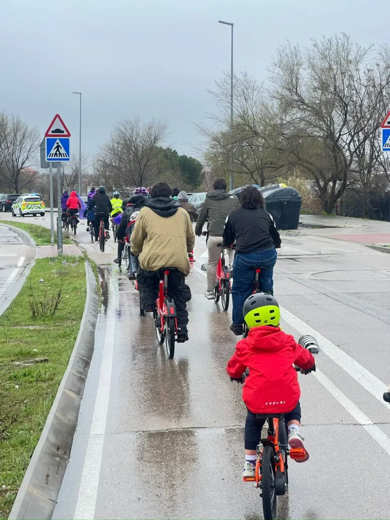 Un grupo de ciclistas, entre los que se encuentran niños y adultos, circulan en bicicleta por un carril bici mojado en Rivas durante la Bicicletada 7M. Las señales de tráfico y los coches aparcados se ven al fondo en este día nublado.