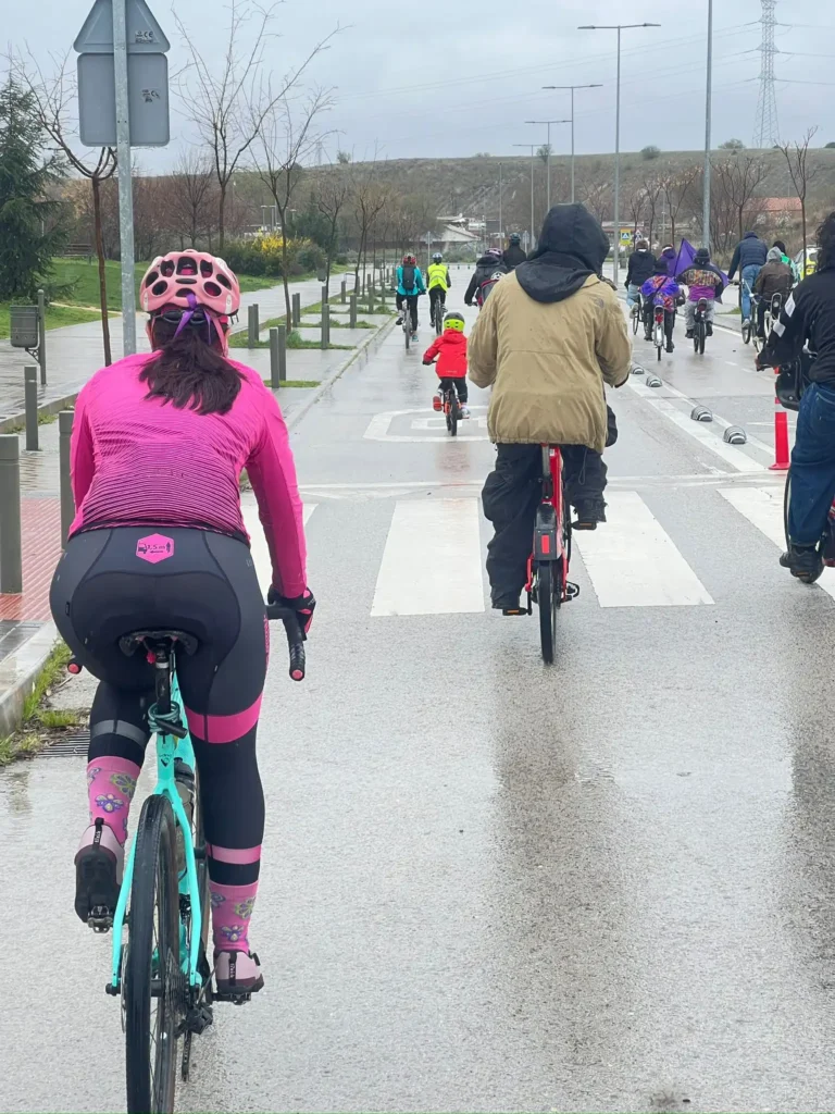 Un grupo de ciclistas circula en bicicleta por una carretera asfaltada y mojada de Rivas. El cielo está nublado, los árboles y las farolas bordean la ruta, y una mujer vestida de ciclista rosa y un hombre con chaqueta morena lideran la Bicicletada 7M.