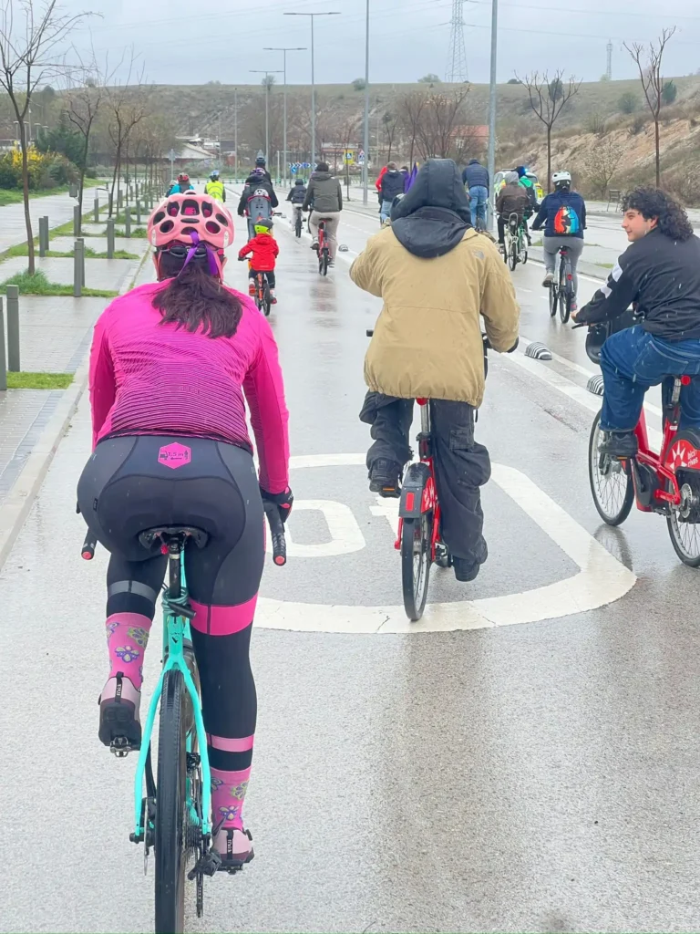 Un grupo de ciclistas circula en bicicleta por una carretera mojada durante la Bicicletada 7M de Rivas. El ciclista más cercano a la cámara lleva chaqueta y casco rosas. La carretera está señalizada con el símbolo de la bicicleta y el cielo está nublado.