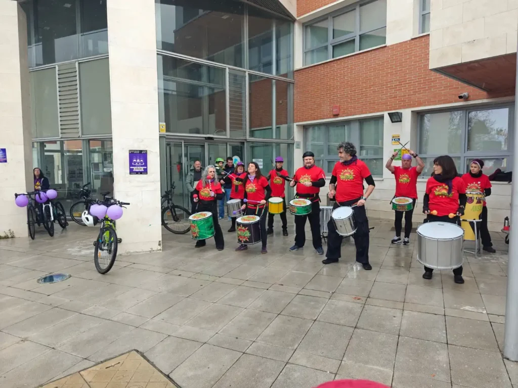 Un grupo de personas con camisetas rojas a juego tocan tambores frente a un moderno edificio durante la Bicicletada 7M en Rivas. Las bicicletas de los ciclistas, decoradas con globos morados, están aparcadas cerca. El suelo mojado sugiere que ha llovido recientemente.