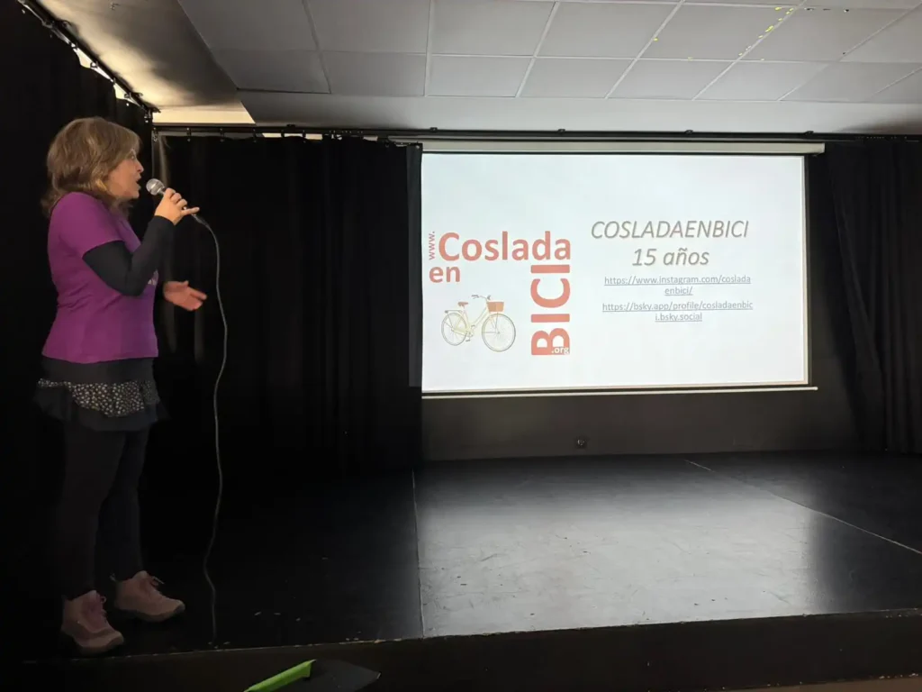 Una mujer habla por un micrófono en un escenario oscuro cerca de una pantalla de proyección que muestra una presentación de COSLADAENBICI 15 años, con enlaces, un gráfico de bicicletas e información para ciclistas sobre la próxima Bicicletada 7M en Rivas.