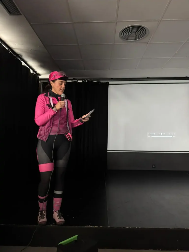 Una persona vestida con ropa deportiva rosa, gorra incluida, sostiene un micrófono y un trozo de papel mientras está de pie en un escenario frente a una pantalla de proyección en blanco en el evento Bicicletada 7M en Rivas, rodeado de ciclistas.
