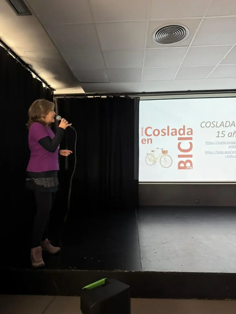 Una mujer de pie en un escenario sosteniendo un micrófono, hablando frente a una pantalla que muestra Coslada en Bici y un gráfico de bicicleta, invitando a los ciclistas a unirse al próximo evento Bicicletada 7M de Rivas con enlaces web mostrados.