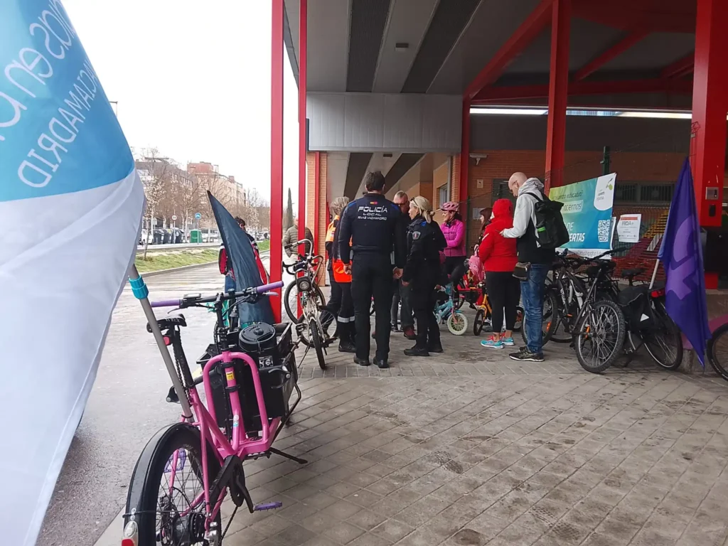 Un grupo de ciclistas con bicicletas se reúnen bajo el voladizo de un edificio cerca de una acera en Rivas. Un agente de policía les acompaña, y en primer plano, a la izquierda, se ve una bandera azul y blanca que señala la Bicicletada 7M.