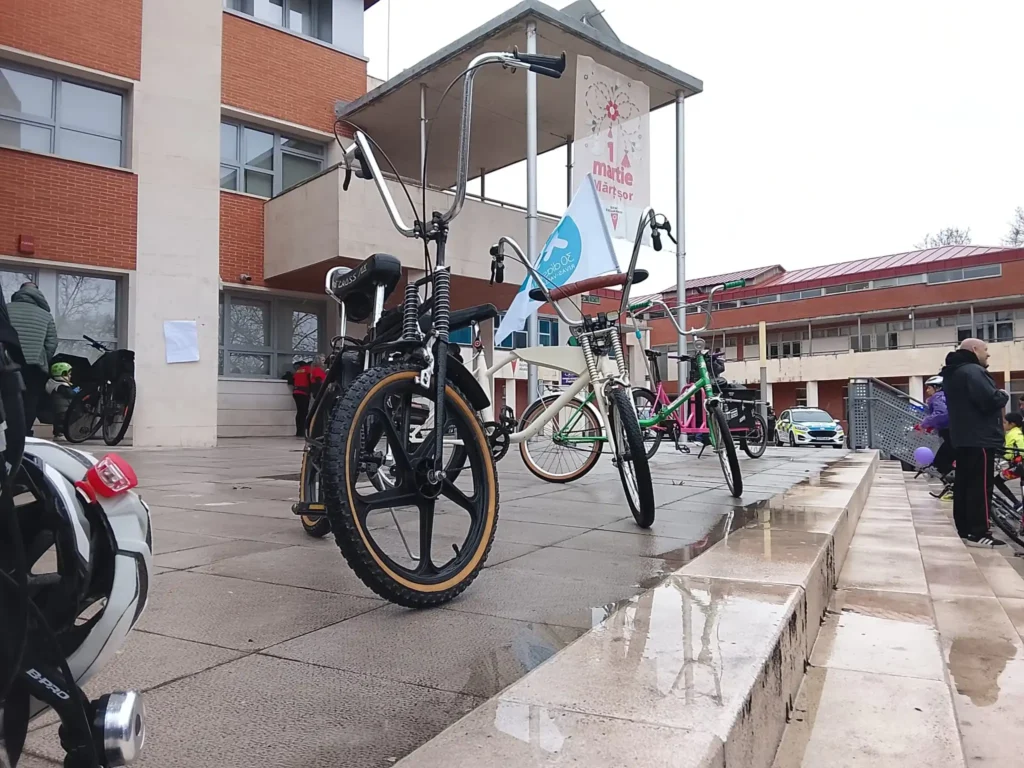 Tres bicicletas con cuadros característicos y grandes manillares están aparcadas en una zona pavimentada y húmeda frente a un moderno edificio durante la Bicicletada 7M. Al fondo se ven ciclistas y otras bicicletas, con una pancarta colgando.