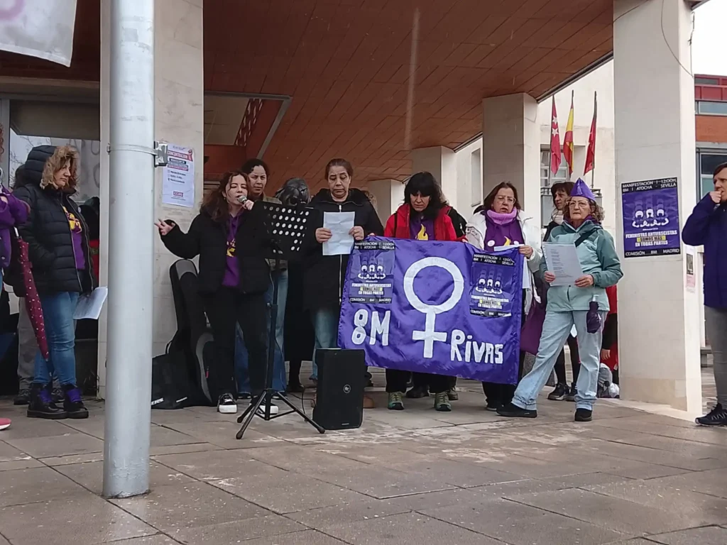Un grupo de personas, entre las que hay ciclistas, permanecen en el exterior de un edificio sosteniendo una pancarta morada con un símbolo feminista y el texto 8M Rivas. Algunos hablan con micrófonos y otros sostienen papeles. Detrás de ellos se ven carteles en las paredes.