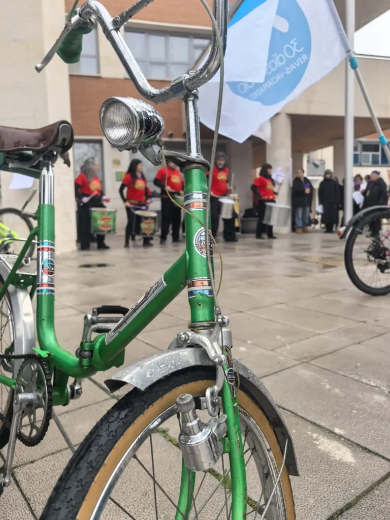 Primer plano de una bicicleta verde de época aparcada sobre el pavimento mojado durante la Bicicletada 7M en Rivas, con ciclistas vestidos de rojo y amarillo cerca. Una bandera azul y blanca ondea detrás del grupo.