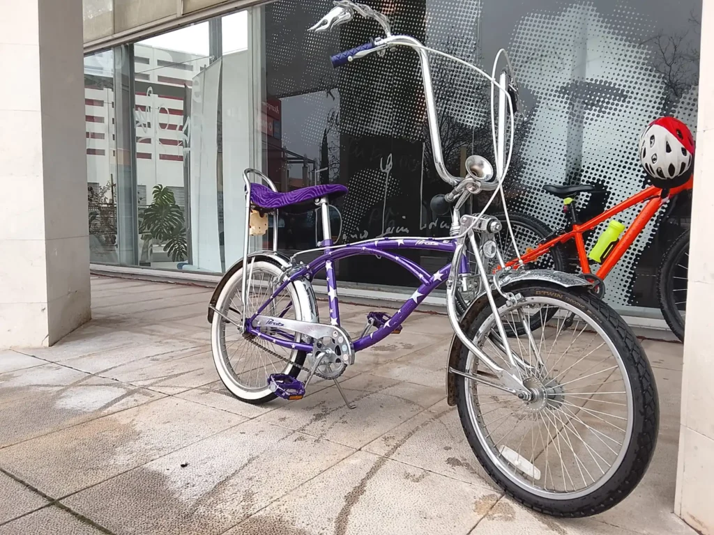 Una bicicleta morada de estilo vintage con manillar alto y asiento de banana está aparcada en una acera junto a otra naranja. Al fondo, cascos de ciclistas aluden a un reciente evento de la Bicicletada 7M en Rivas junto a la fachada acristalada del edificio.