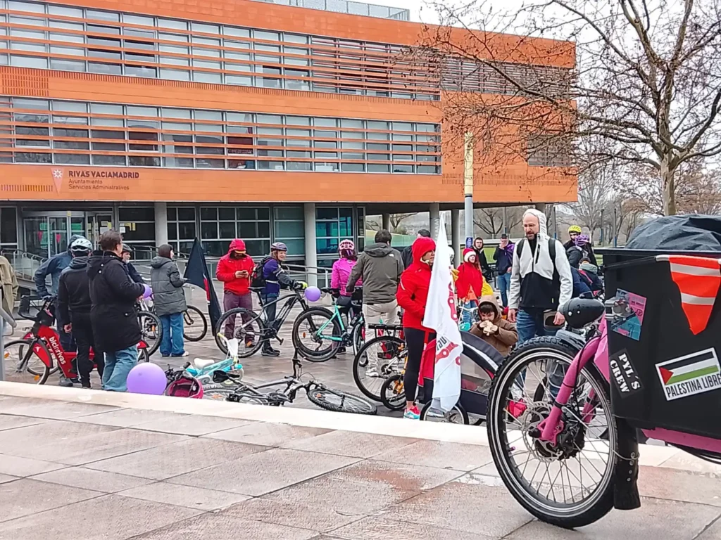 Un grupo de ciclistas se reúne al aire libre en Rivas para la Bicicletada 7M, frente a un edificio naranja. Algunos sostienen banderas, unos pocos llevan casco y un adulto tira de un niño en un remolque de bicicleta. Entre el grupo se ven globos y pancartas.