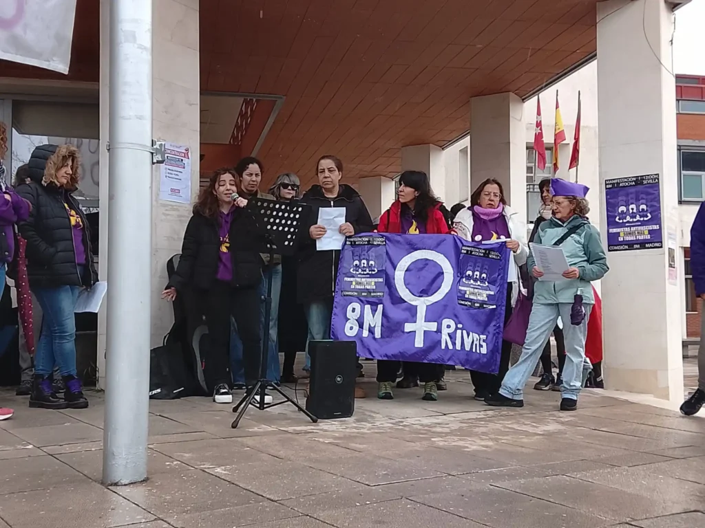 Un grupo de personas, entre ellas ciclistas de Rivas, permanecen de pie frente a un edificio. Algunos sostienen una pancarta morada con un símbolo femenino y "8M Rivas", mientras otros leen en papeles cerca de un micrófono.
