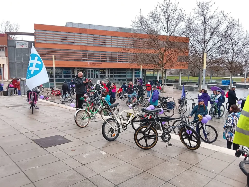 Un grupo de ciclistas, niños y adultos, se reúnen con bicicletas en una plaza abierta cerca de un moderno edificio durante la Bicicletada 7M en Rivas. Algunas bicicletas llevan globos atados y las banderas ondean mientras al fondo se ven árboles sin hojas.