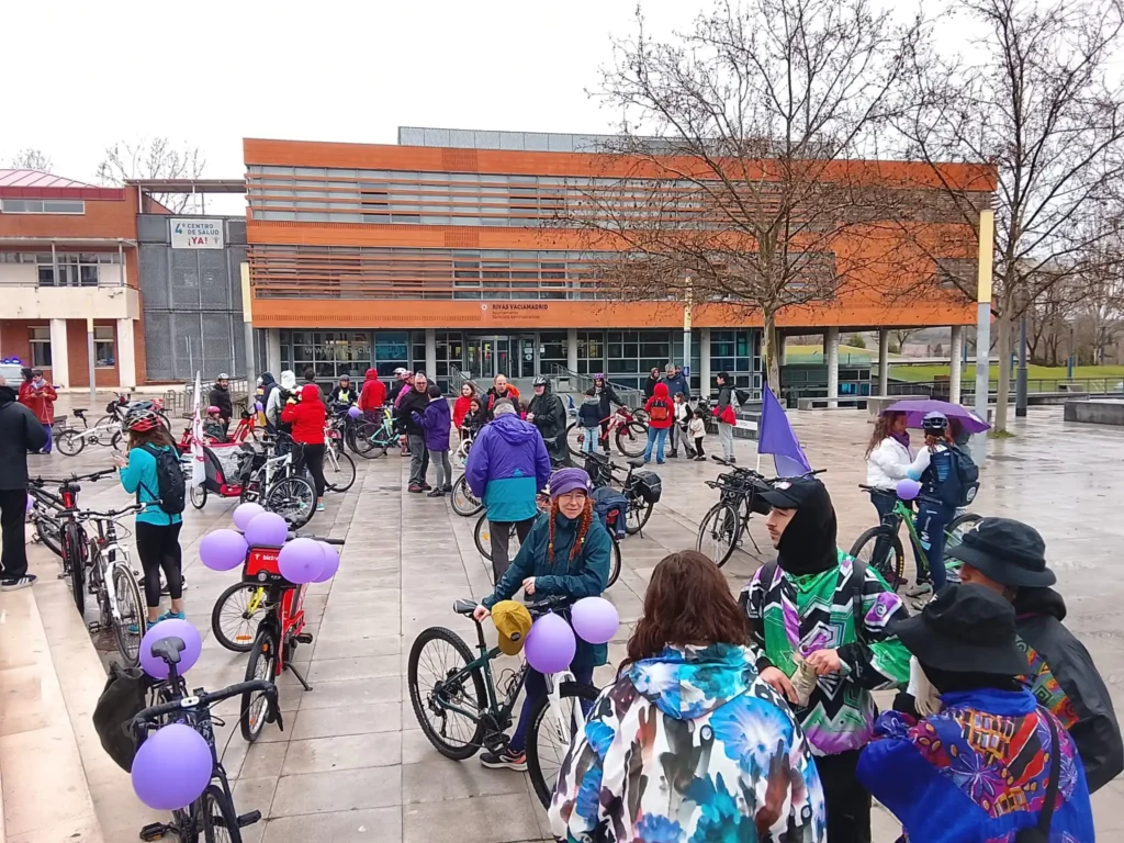 Ciclistas se reúnen para la Bicicletada 7M en Rivas, con bicicletas adornadas con globos morados. Están en una plaza pavimentada ante un moderno edificio en un día nublado, abrigados con chaquetas y sombreros. Árboles desnudos enmarcan la escena.