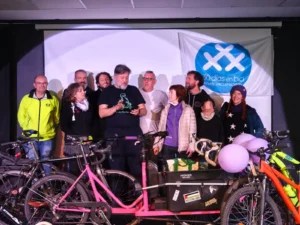 Un grupo de ciclistas de pie en un escenario en Rivas detr&aacute;s de varias bicicletas y material ciclista, con una pancarta de "30 d&iacute;as en bici" en la pared. Los individuos parecen estar escuchando al hombre de delante que lee en un papel.