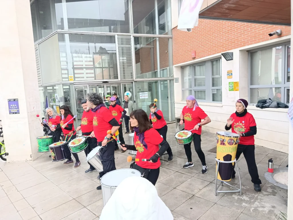 Un grupo de personas con camisetas rojas a juego con dibujos amarillos tocan tambores e instrumentos de percusión en el exterior de un moderno edificio de Rivas, al paso de los ciclistas de la Bicicletada 7M.
