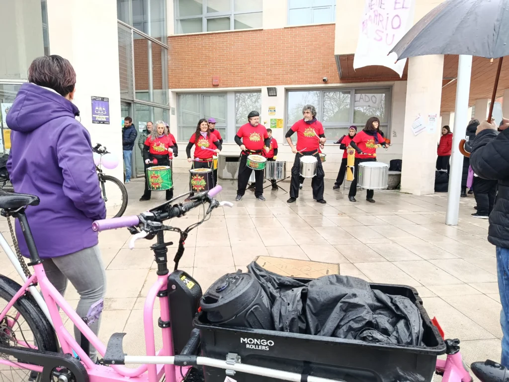 Un grupo de personas con camisetas rojas tocan tambores en el exterior de un edificio durante la Bicicletada 7M en Rivas. En primer plano, una bicicleta rosa con una caja de carga negra está aparcada. Varios ciclistas y espectadores observan, algunos con paraguas en la mano.