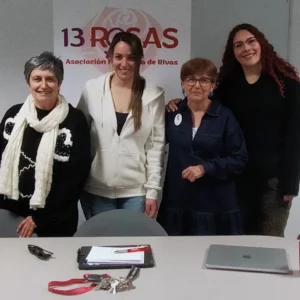 Cuatro mujeres de la nueva Junta Directiva de la Asociación Feminista 13Rosas están juntas delante de una pancarta de 13Rosas. Sonriendo a la cámara, están reunidas detrás de una mesa con un portapapeles, llaves, un cordón y un ordenador portátil.